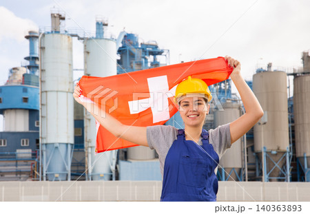 Young female engineer in helmet waving state flag of Switzerland while standing in front of big tanks at chemical plant Young female engineer in helmet waving state flag of Switzerland while standing in front of big tanks at chemical plant 140363893