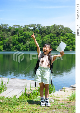 新緑の湖畔でしおりを持ち元気に手を挙げる遠足の女子小学生 140363971