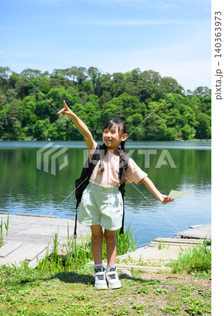 新緑の湖畔でしおりを持ち元気に手を挙げる遠足の女子小学生 140363973