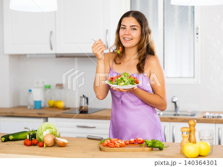 Portrait of girl in purple nightgown tasting vegetable salad at kitchen Portrait of girl in purple nightgown tasting vegetable salad at kitchen 140364301
