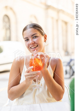 Smiling stylish young woman in light dress drinking cocktail sitting at outdoor cafe Smiling stylish young woman in light dress drinking cocktail sitting at outdoor cafe 140364449