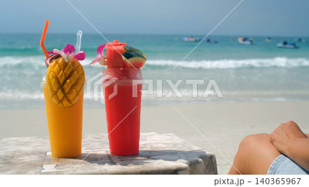 Young woman relaxing in a beach lounge chair next to a table with mango and watermelon smoothies with sea and beach in background. Beach leisure concept Young woman relaxing in a beach lounge chair next to a table with mango and watermelon smoothies with sea and beach in background. Beach leisure concept 140365967