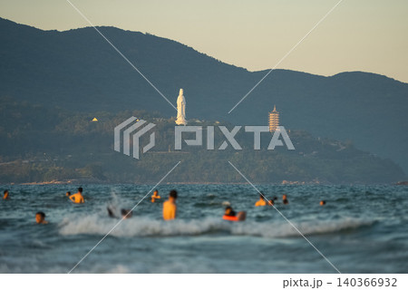 People swim in Danang sea, Lady Buddha statue and Linh Ung Pagoda above waves and tourists in Da Nang, Coastal view of Son Tra Peninsula with Buddhist landmark, mountains and ocean shoreline People swim in Danang sea, Lady Buddha statue and Linh Ung Pagoda above waves and tourists in Da Nang, Coastal view of Son Tra Peninsula with Buddhist landmark, mountains and ocean shoreline 140366932