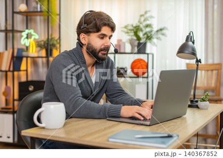 Indian man with headset and mic at laptop polite support agent listens and writes notes in notebook Indian man with headset and mic at laptop polite support agent listens and writes notes in notebook 140367852