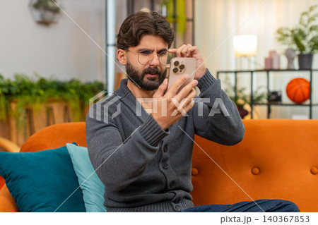 Indian man with eyeglasses squints at smartphone screen tries to read small text poor vision focus Indian man with eyeglasses squints at smartphone screen tries to read small text poor vision focus 140367853