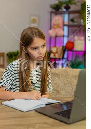 Child girl kid doing online homework on laptop and writing math notes sitting at home table Child girl kid doing online homework on laptop and writing math notes sitting at home table 140368019