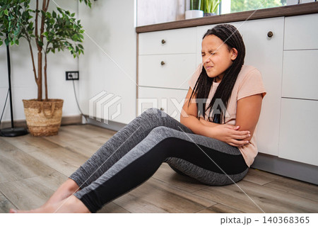 Girl experiencing stomach pain sitting on kitchen floor Girl experiencing stomach pain sitting on kitchen floor 140368365
