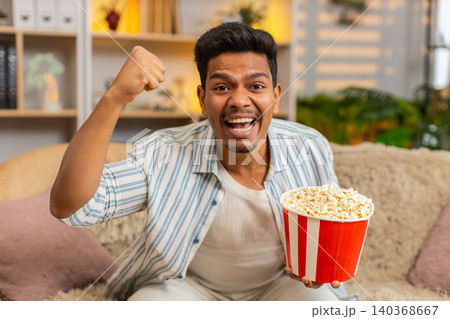 Indian man on home sofa eating popcorn and cheering for sports team with joyful win reaction indoors Indian man on home sofa eating popcorn and cheering for sports team with joyful win reaction indoors 140368667