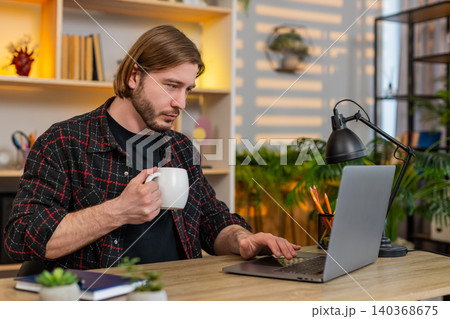 Young man on home table drinking coffee while reading laptop screen during remote work break at home 140368675