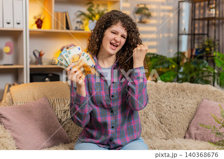 Young woman on home sofa counting cash and smiling after salary bonus win or debt repayment indoors Young woman on home sofa counting cash and smiling after salary bonus win or debt repayment indoors 140368676