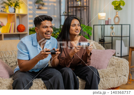 Indian couple man and woman sitting on home sofa playing video games with wireless gamepads at home Indian couple man and woman sitting on home sofa playing video games with wireless gamepads at home 140368737