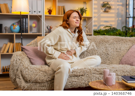 Young woman on home sofa holding stomach in pain from menstrual cramps and female health discomfort 140368746