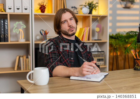 Young man on home table writing notes in notebook while planning tasks for tomorrow with calm focus Young man on home table writing notes in notebook while planning tasks for tomorrow with calm focus 140368851