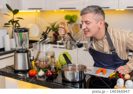 Middle-aged man tasting homemade vegetable soup in kitchen while checking flavor for healthy lunch 140368947