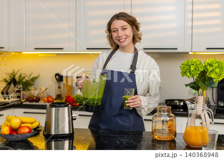 Young woman preparing fresh green smoothie in kitchen at home for healthy breakfast wellness drink 140368948