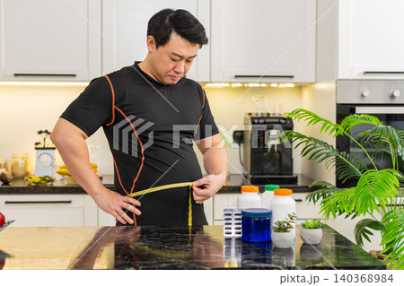 Heavy Asian adult man measures waist with tape upset about big belly fat weight gain at home kitchen Heavy Asian adult man measures waist with tape upset about big belly fat weight gain at home kitchen 140368984