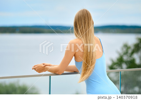 Back view of woman with long blond hair standing on balcony and gazes out at water Back view of woman with long blond hair standing on balcony and gazes out at water 140370918