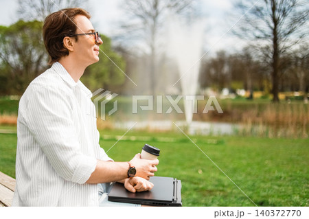 A man working as a freelancer sits on a park bench with a laptop and coffee, enjoying remote work outdoors near a fountain. A man working as a freelancer sits on a park bench with a laptop and coffee, enjoying remote work outdoors near a fountain. 140372770