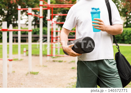 Fit man standing at an outdoor workout park with a foam roller and a shaker bottle, preparing for a fitness training session and active lifestyle routine. Fit man standing at an outdoor workout park with a foam roller and a shaker bottle, preparing for a fitness training session and active lifestyle routine. 140372771