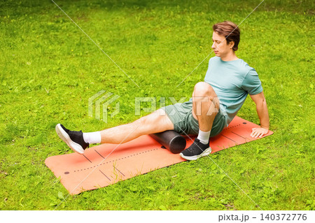 A man uses a foam roller on a mat for myofascial release and post-workout recovery, targeting leg muscle tension with self-massage outdoors in a park. A man uses a foam roller on a mat for myofascial release and post-workout recovery, targeting leg muscle tension with self-massage outdoors in a park. 140372776