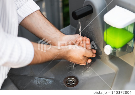 Close-up of a man washing his hands under running water at an outdoor portable sink with a green liquid soap dispenser, focusing on hygiene and sanitation. Close-up of a man washing his hands under running water at an outdoor portable sink with a green liquid soap dispenser, focusing on hygiene and sanitation. 140372804
