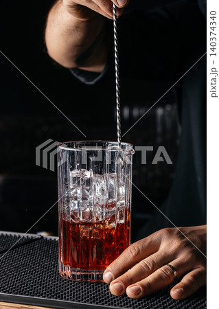 Bartender stirring red cocktail with ice in crystal mixing glass at dark bar 140374340