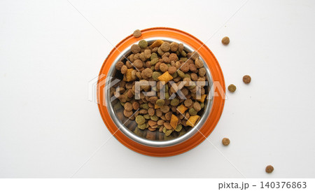 Pet food grains. Dry animal food bits in an orange metal bowl on a white studio background. Cat or dog kibble heap. Creative funny image for ad of pet products. Top view. Carnivore of domestic animals Pet food grains. Dry animal food bits in an orange metal bowl on a white studio background. Cat or dog kibble heap. Creative funny image for ad of pet products. Top view. Carnivore of domestic animals 140376863