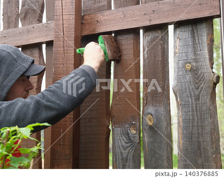 Brown oil for protection of wood by staining. Painting fence for protecting and pests. Woodworker applying finish to pine deck board. Repairman varnishing with brush wooden fencing wall plank surface. Brown oil for protection of wood by staining. Painting fence for protecting and pests. Woodworker applying finish to pine deck board. Repairman varnishing with brush wooden fencing wall plank surface. 140376885