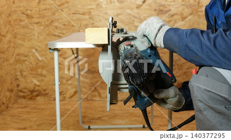 Builder saws a board with a circular saw cutting a wooden plank in a joinery. Carpenter uses portable circular saw tool to cut wood at wooden table in workshop. Worker sawing pine board. Real image Builder saws a board with a circular saw cutting a wooden plank in a joinery. Carpenter uses portable circular saw tool to cut wood at wooden table in workshop. Worker sawing pine board. Real image 140377295