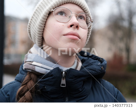 Teenager Girl Walking Alone on City Street. Portrait of 14 years old girl wearing eyeglasses, wool hat and winter jacket walks around the city. Urban Style. Rainy mood weather. Cold season. One person 140377313