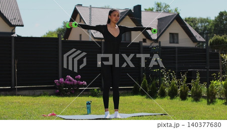 Young beautiful sports girl in leggings does exercises with dumbbells. Healthy lifestyle. Woman goes in for sports outdoor at home. 140377680