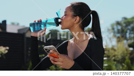 Young sport woman resting and using smartphone, drink water after workout in backyard. Sports female doing outdoor training. Young sport woman resting and using smartphone, drink water after workout in backyard. Sports female doing outdoor training. 140377707