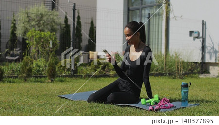Young sport woman resting and using smartphone, after workout in backyard. Mobile application for training. Sports female doing outdoor training. 140377895