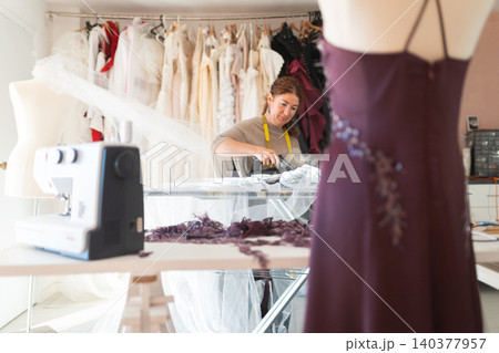 Craftswoman designer tailor working on a sewing machine with a purple dress on a mannequin and various fabrics hanging in the background of a bright studio Craftswoman designer tailor working on a sewing machine with a purple dress on a mannequin and various fabrics hanging in the background of a bright studio 140377957
