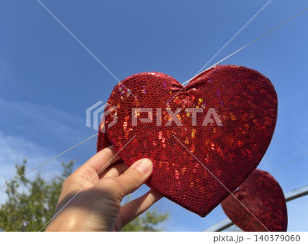 a hand holding a large, shimmering red heart against a vibrant blue sky. 140379060