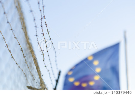 Barbed wire fence and European Union flag near border area under clear sky Barbed wire fence and European Union flag near border area under clear sky 140379440