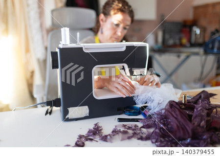Woman sewing with a machine in a workspace filled with fabric and sewing tools during the day Woman sewing with a machine in a workspace filled with fabric and sewing tools during the day 140379455