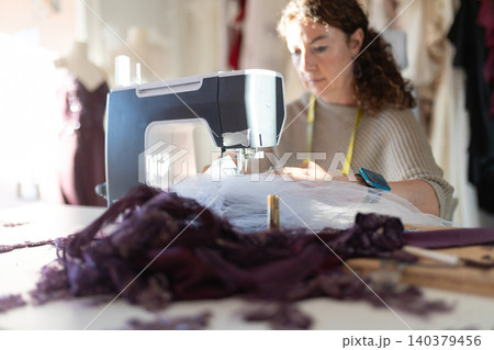 Woman sewing fabric at a table with a sewing machine in a workshop during daytime hours Woman sewing fabric at a table with a sewing machine in a workshop during daytime hours 140379456