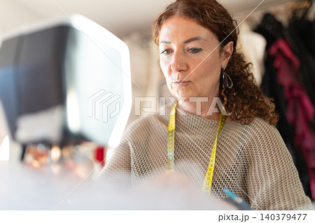 Woman sewing at a sewing machine in a workshop during daytime while creating clothing items and adjusting fabric pieces Woman sewing at a sewing machine in a workshop during daytime while creating clothing items and adjusting fabric pieces 140379477