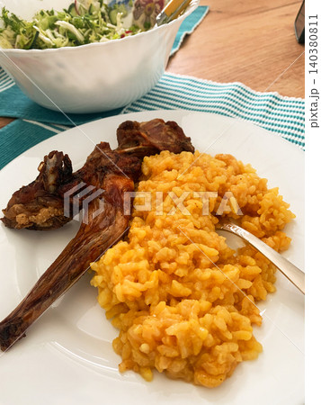A meal is served on a white plate, showing rice colored with turmeric and pieces of baked rabbit beside a fresh salad in a bowl. The meal is set on a wooden table with a blue cloth 140380811