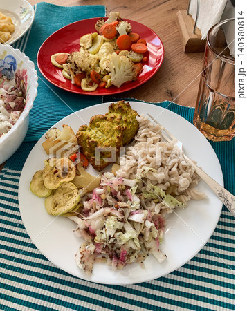 A plate holds gluten-free pasta, broccoli fritters, baked vegetables, and a fresh salad. Another red plate has more baked vegetables with cherry tomatoes A plate holds gluten-free pasta, broccoli fritters, baked vegetables, and a fresh salad. Another red plate has more baked vegetables with cherry tomatoes 140380814