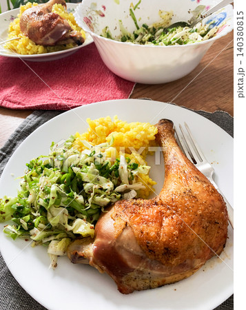 A meal features turmeric rice next to a baked chicken leg and a fresh salad on a white plate placed on a wooden table. The setting is warm and inviting A meal features turmeric rice next to a baked chicken leg and a fresh salad on a white plate placed on a wooden table. The setting is warm and inviting 140380815