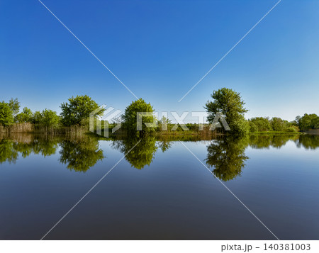 Serene lakeside landscape with trees reflecting on calm water under a clear blue sky Serene lakeside landscape with trees reflecting on calm water under a clear blue sky 140381003