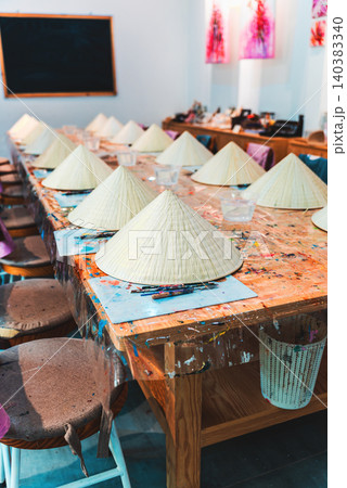 Non la hat painting workshop with brushes palettes and colorful paints on wooden table, Traditional Vietnam craft activity painting conical hats in creative art studio setting Non la hat painting workshop with brushes palettes and colorful paints on wooden table, Traditional Vietnam craft activity painting conical hats in creative art studio setting 140383340