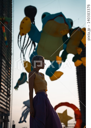 young woman walking on sand beach at colorful kite toys festival during summer sunset, girl enjoying freedom and joy at outdoor event by sea 140383376