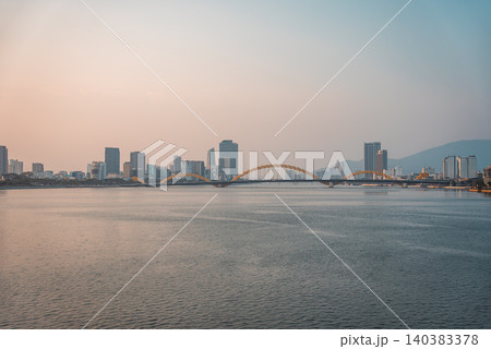 Dragon bridge over calm Han river at sunset with soft golden light and peaceful city skyline of Da Nang, tranquil urban landscape with serene water and warm evening atmosphere, Danang Vietnam 140383378