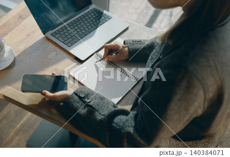Asian young adult writing in notebook and holding smartphone near laptop in cafe Asian young adult writing in notebook and holding smartphone near laptop in cafe 140384071