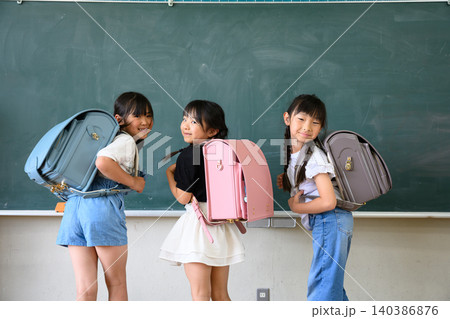 教室の黒板の前でカラフルなランドセルを背負い笑顔で振り返る新入生の女の子たち 140386876