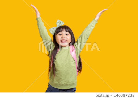A happy Asian schoolgirl raises her hand with her backpack on; against a yellow background, the excited child exudes a sense of success and confidence. A happy Asian schoolgirl raises her hand with her backpack on; against a yellow background, the excited child exudes a sense of success and confidence. 140387238