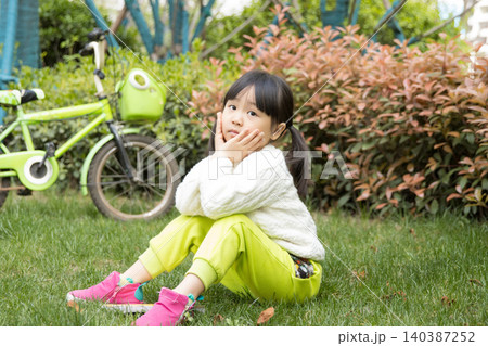 A lovely Asian girl sits on the grass outdoors, resting her chin on her hands, embodying a relaxed outdoor lifestyle. A lovely Asian girl sits on the grass outdoors, resting her chin on her hands, embodying a relaxed outdoor lifestyle. 140387252
